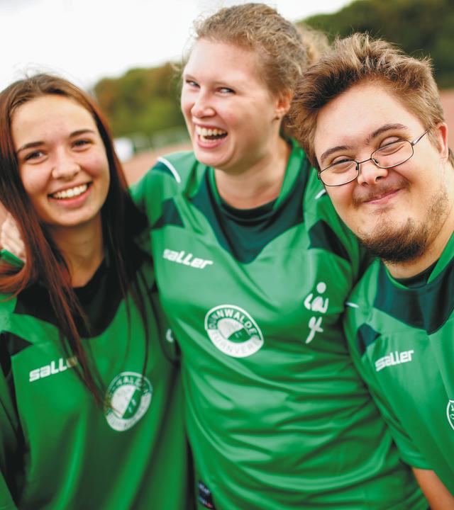 Drei junge Menschen im Sportdress Arm in Arm, darunter ein junger Mann mit Trisomie 21