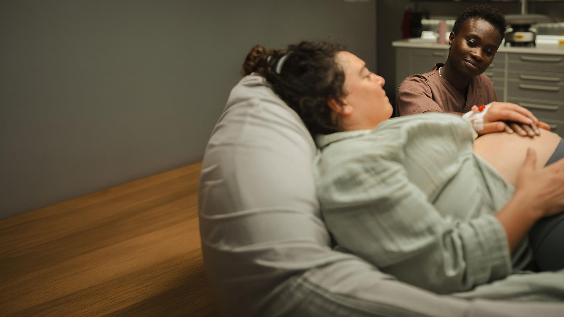 "PUSH - Akzeptanz": Sibel (Nuriye Jendroßek) liegt im Patientenbett und schaut zu Tara (Lorna Ishema), die an ihrem Bett sitzt und eine Hand auf Sibels Babybauch legt. Sibel hält Taras Hand.