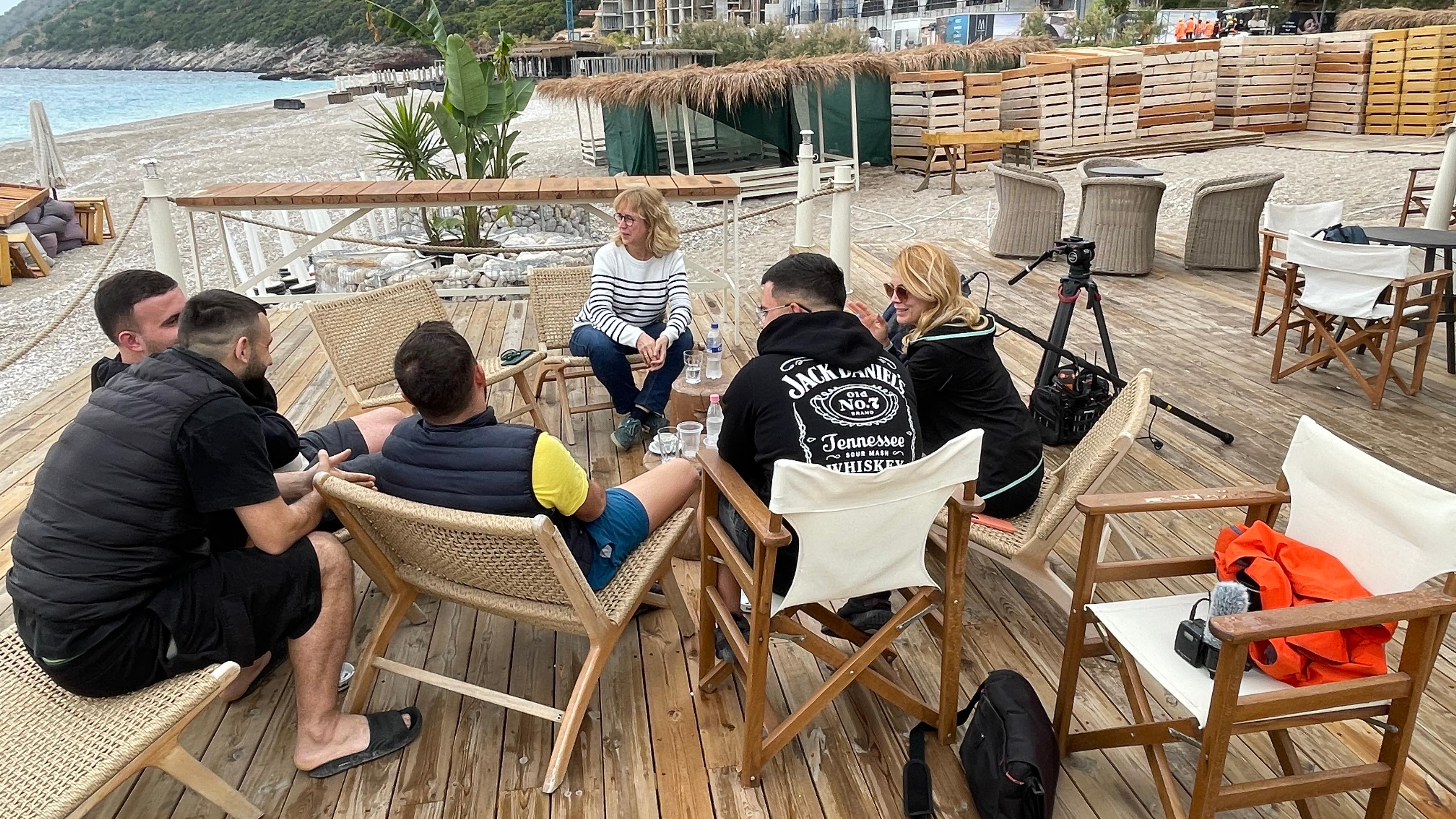 Vier junge Männer und eine Frau sitzen auf Klappstühlen im Halbkreis. Sie sprechen mit einer blonden Frau. Die Gruppe sitzt auf einer großen Terasse am Strand. Im Hintergrund ist die grüne Küste und das Meer zu sehen