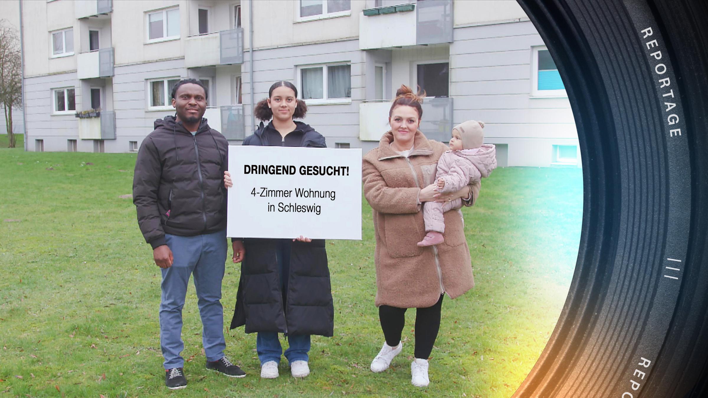 Steven-Ken und Tatyana stehen mit ihren Kindern auf einer Wiese vor einem Wohnblock. Die große Tochter hält ein großes Schild in die Kamera: "Dringend gesucht! 4-Zimmer-Wohnung in Schleswig".
