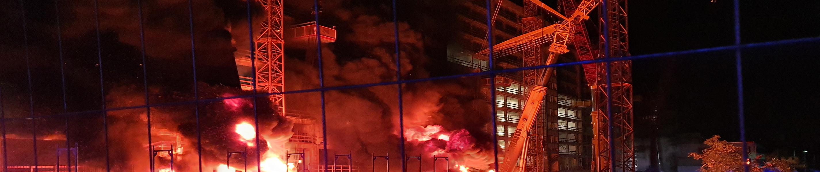 "Albtraum Wohnen": Blick durch den Bauzaun auf eine nächtliche Baustelle. An zwei Stellen stehen Maschinen in Brand.