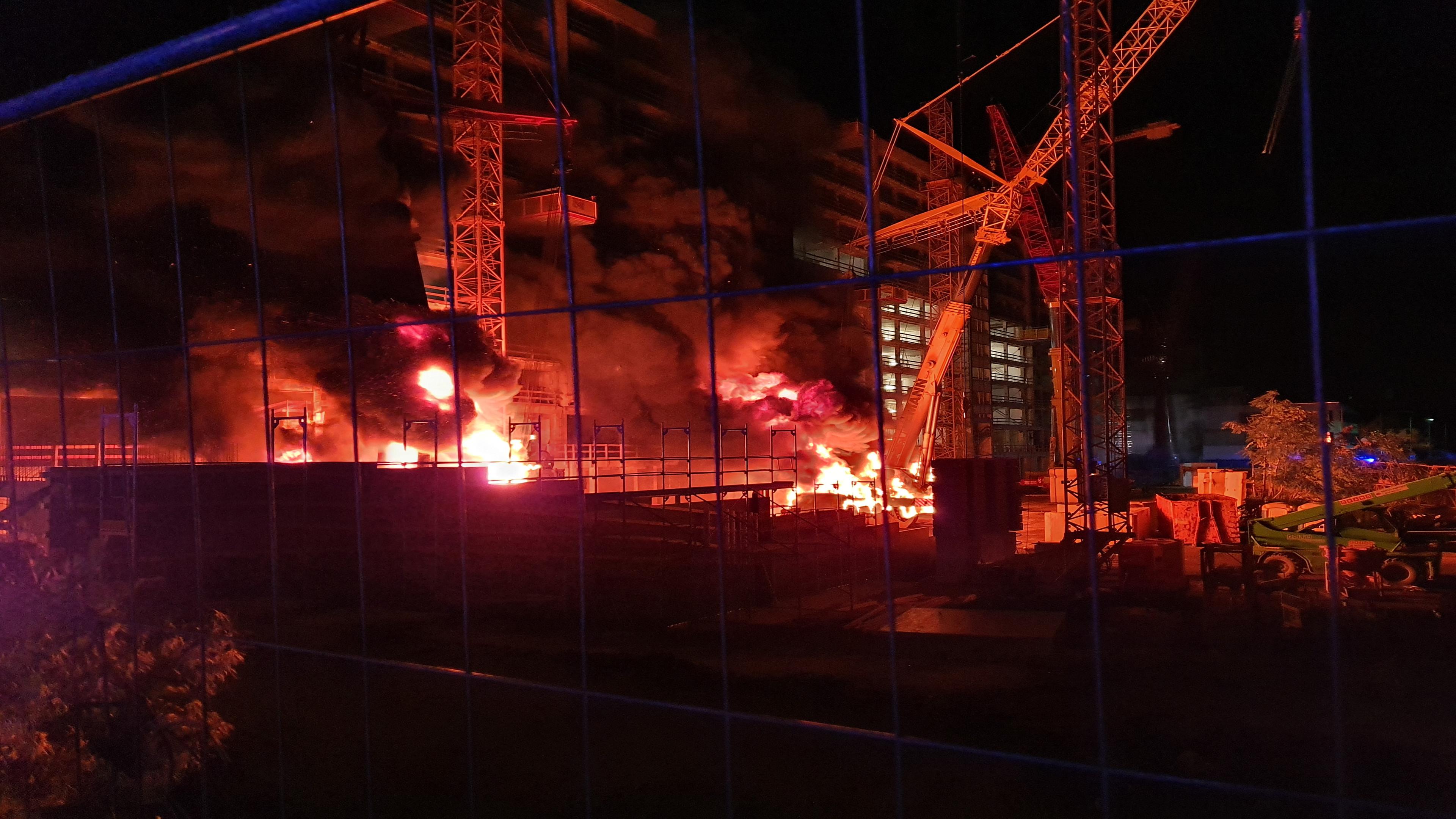 "Albtraum Wohnen": Blick durch den Bauzaun auf eine nächtliche Baustelle. An zwei Stellen stehen Maschinen in Brand.