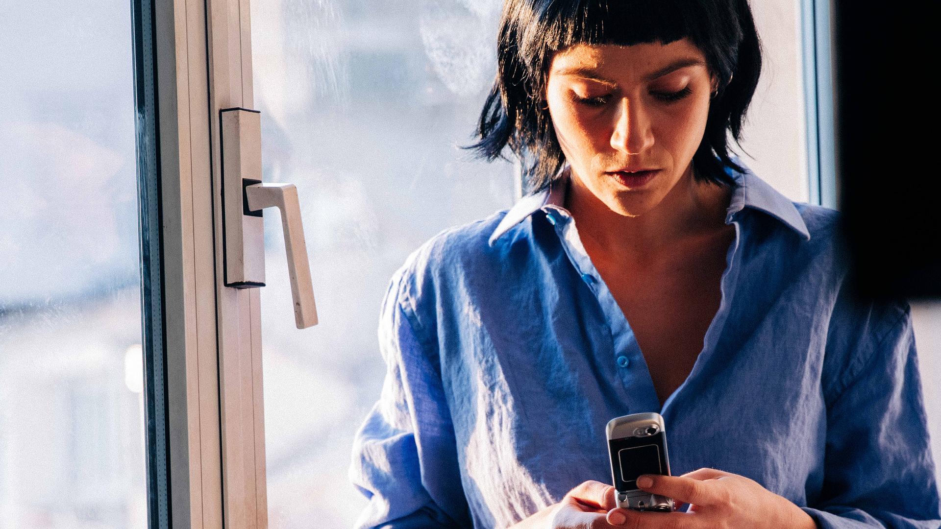 "Survivors": Alex (Sophie Pfennigstorf) steht in ihrer Wohnung vor einer Fensterfront und liest eine Nachricht auf ihrem Handy.