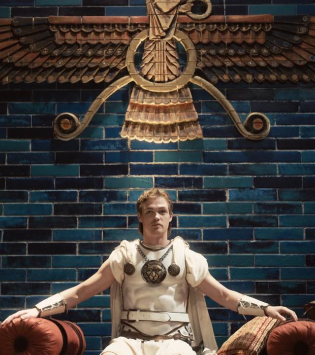 Alexander der Große sitzt als junger Mann auf einem Thron.