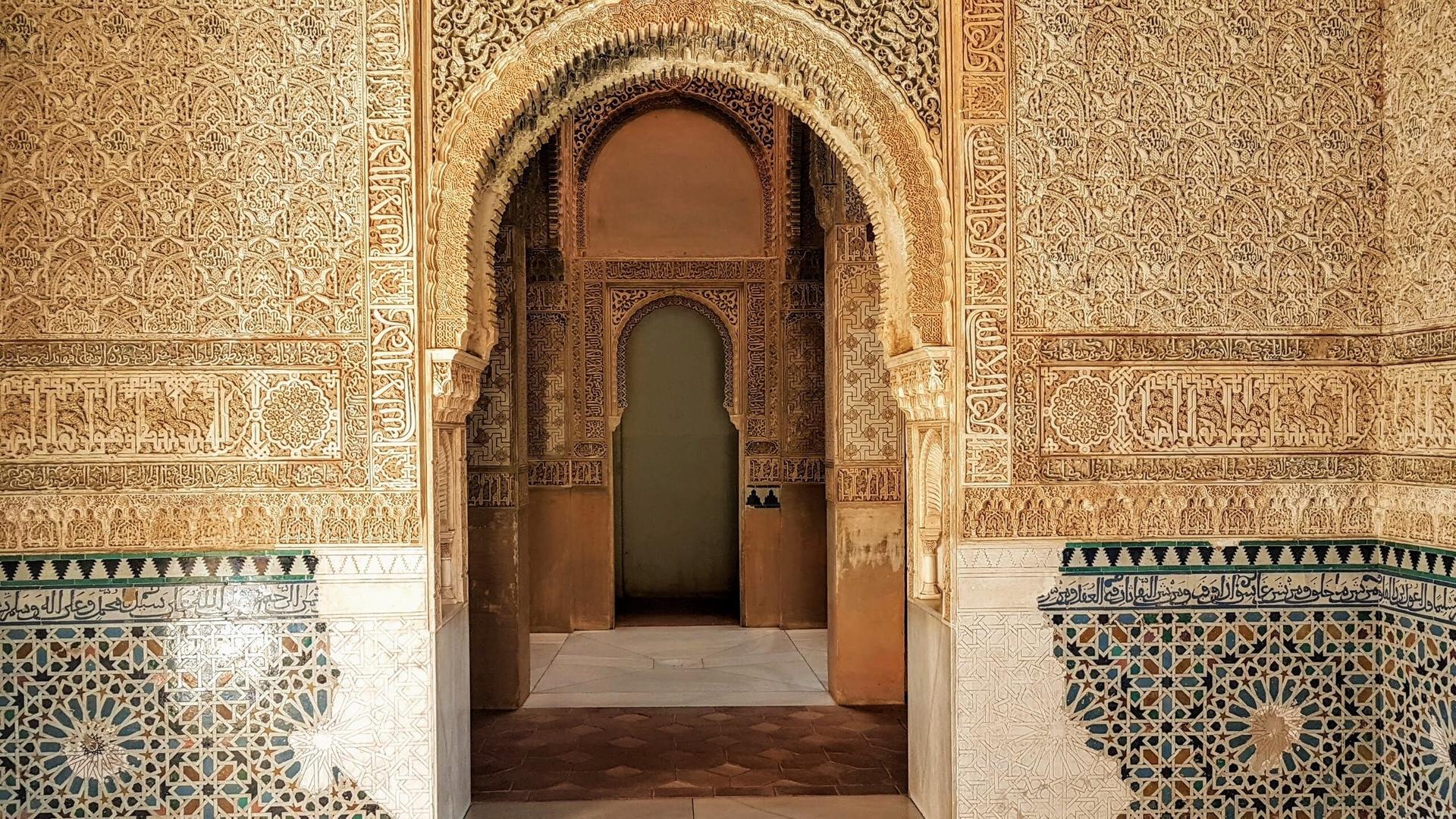 Das Bild zeigt einen Innenraum der Alhambra, einer historischen Festung in Granada, Spanien. Die Wände sind reich verziert mit feinen, detailreichen Ornamenten und Schriftzügen, die typisch für die maurische Architektur sind.   Im Vordergrund erkennt man einen großen, geschwungenen Bogen, der in einen weiteren Raum führt. Die Wände sind beige mit dekorativen Elementen in verschiedenen Brauntönen, die kunstvoll geschnitzt sind.   An der unteren Wand ist ein Mosaik mit geometrischen Mustern in blau, grün und weiß zu sehen. Der Boden besteht aus großen, hellen Steinfliesen und einem speziellen Muster. Die gesamte Szenerie strahlt eine Atmosphäre von Eleganz und historischer Bedeutung aus. Licht gelangt durch die Bögen und schafft einen warmen, einladenden Raum.