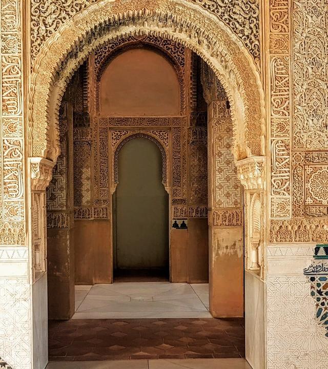 Das Bild zeigt einen Innenraum der Alhambra, einer historischen Festung in Granada, Spanien. Die Wände sind reich verziert mit feinen, detailreichen Ornamenten und Schriftzügen, die typisch für die maurische Architektur sind.   Im Vordergrund erkennt man einen großen, geschwungenen Bogen, der in einen weiteren Raum führt. Die Wände sind beige mit dekorativen Elementen in verschiedenen Brauntönen, die kunstvoll geschnitzt sind.   An der unteren Wand ist ein Mosaik mit geometrischen Mustern in blau, grün und weiß zu sehen. Der Boden besteht aus großen, hellen Steinfliesen und einem speziellen Muster. Die gesamte Szenerie strahlt eine Atmosphäre von Eleganz und historischer Bedeutung aus. Licht gelangt durch die Bögen und schafft einen warmen, einladenden Raum.
