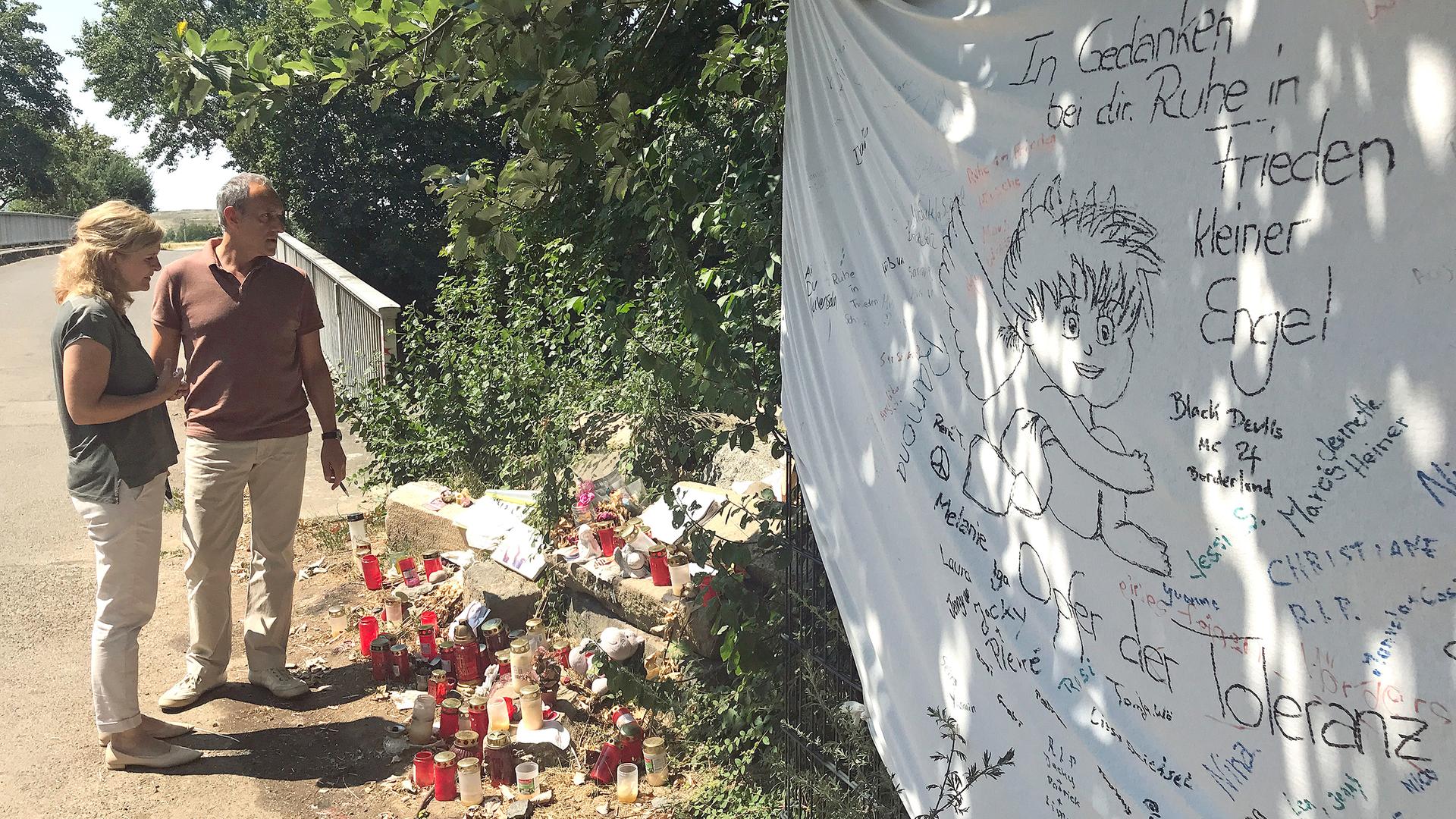 Die beiden Autoren stehen vor einer Brücke und schauen auf eine Erinnerungsstelle mit vielen Kerzen und einem gespannten Tuch, auf dem viele Namen, Zeichnungen und Botschaften stehen.