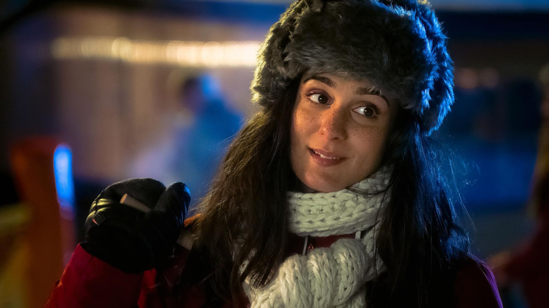 Eine junge Frau mit langen dunklen Haaren steht draußen bei Nacht. Sie trägt eine graue Fellmütze, einen dicken weißen Strickschal und einen roten Wintermantel. In der Hand hält sie einen Skistock, ihr Gesichtsausdruck wirkt nachdenklich und leicht lächelnd.