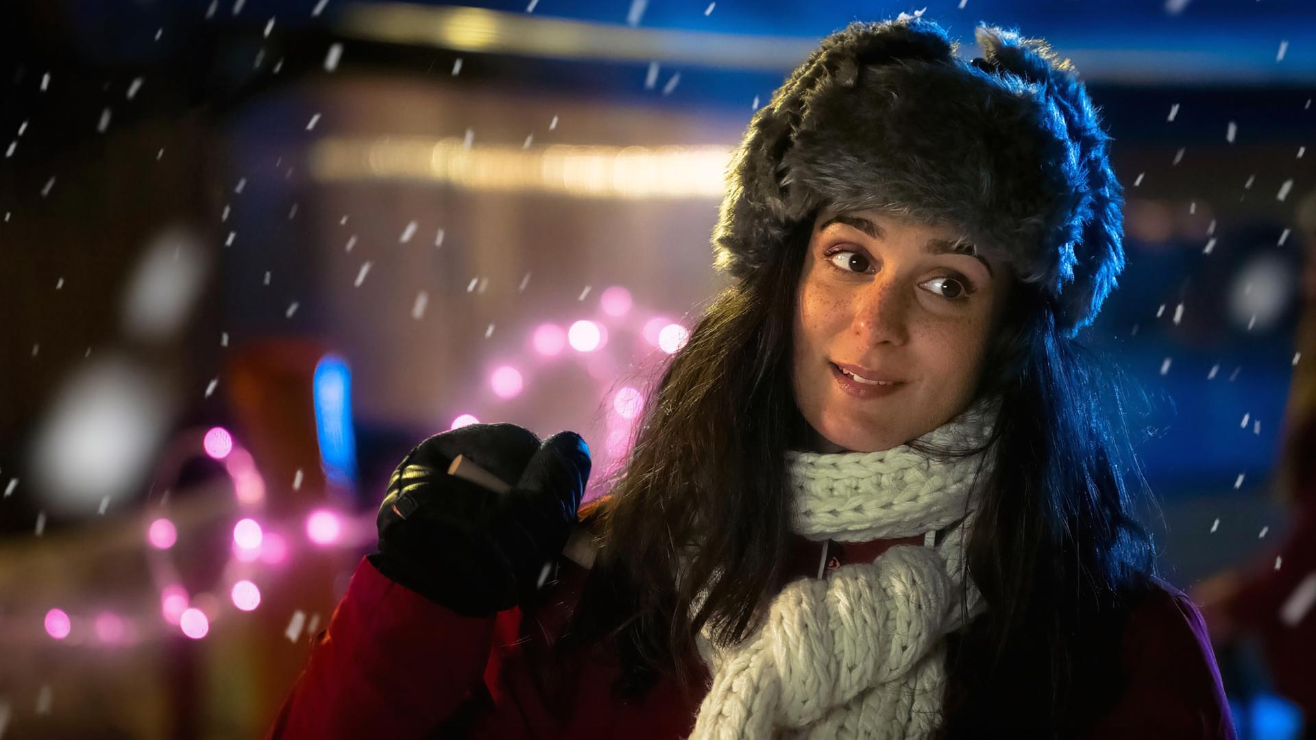 Eine junge Frau mit langen dunklen Haaren steht draußen bei Nacht. Sie trägt eine graue Fellmütze, einen dicken weißen Strickschal und einen roten Wintermantel. In der Hand hält sie einen Skistock, ihr Gesichtsausdruck wirkt nachdenklich und leicht lächelnd.