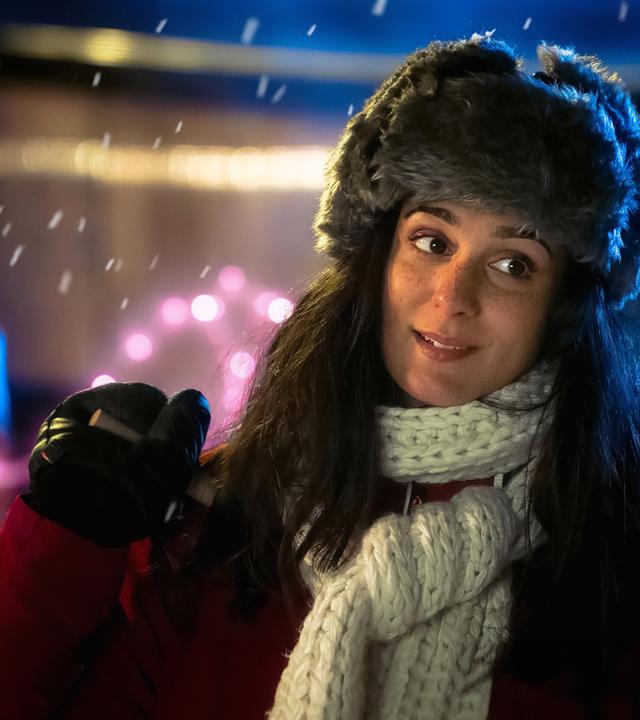 Eine junge Frau mit langen dunklen Haaren steht draußen bei Nacht. Sie trägt eine graue Fellmütze, einen dicken weißen Strickschal und einen roten Wintermantel. In der Hand hält sie einen Skistock, ihr Gesichtsausdruck wirkt nachdenklich und leicht lächelnd.