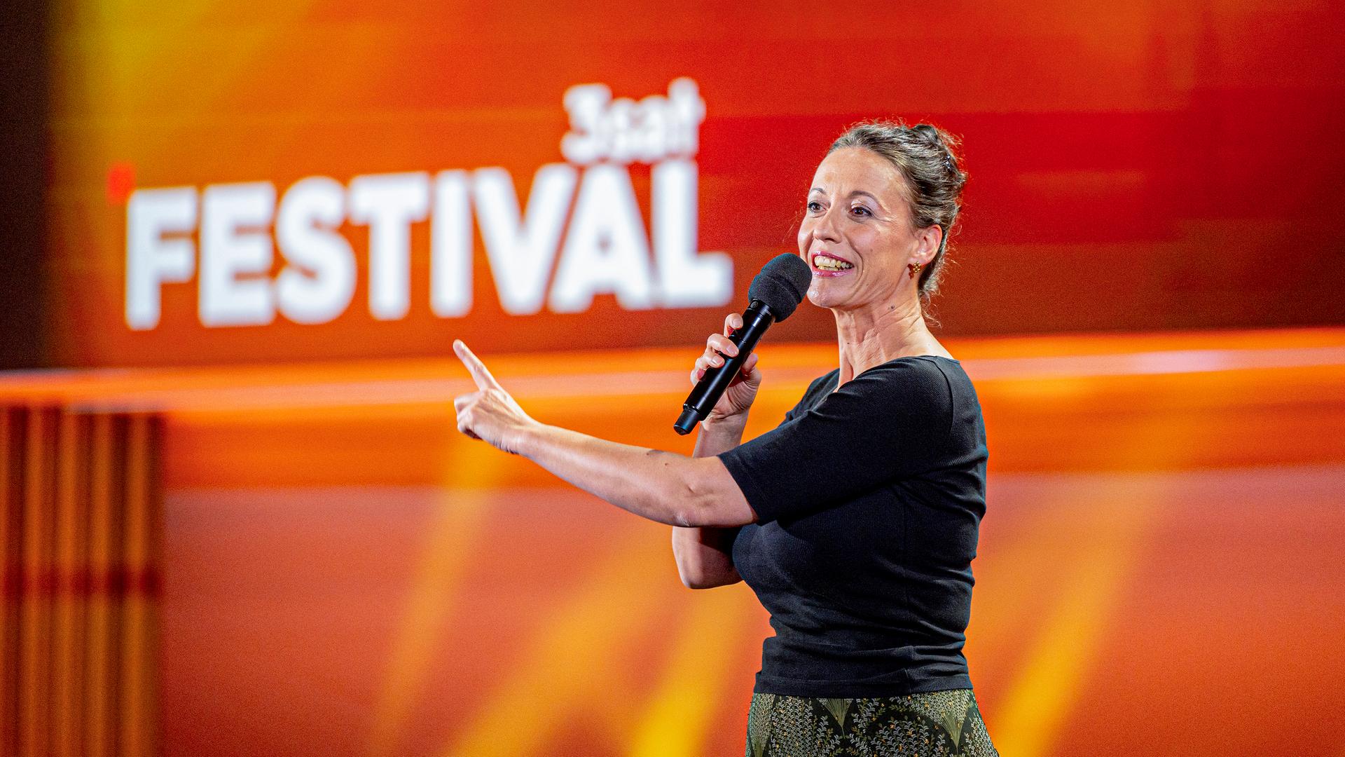 Kabarettistin steht auf der Mitte der Bühne. In ihrer rechten Hand hält sie ein Mikrofon. Mit der linken Hand zeigt sie mit einem Finger nach vorne. Der Hintergrund ist rot und orange beleuchtet.