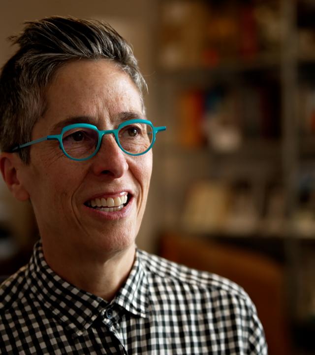 Porträtfoto von Alison Bechdel, einer Frau mit kurzen zurückgekämmten Haaren, die eine auffällige runde türkisfarbene Brille und ein schwarz-weiß-kariertes Hemd trägt.