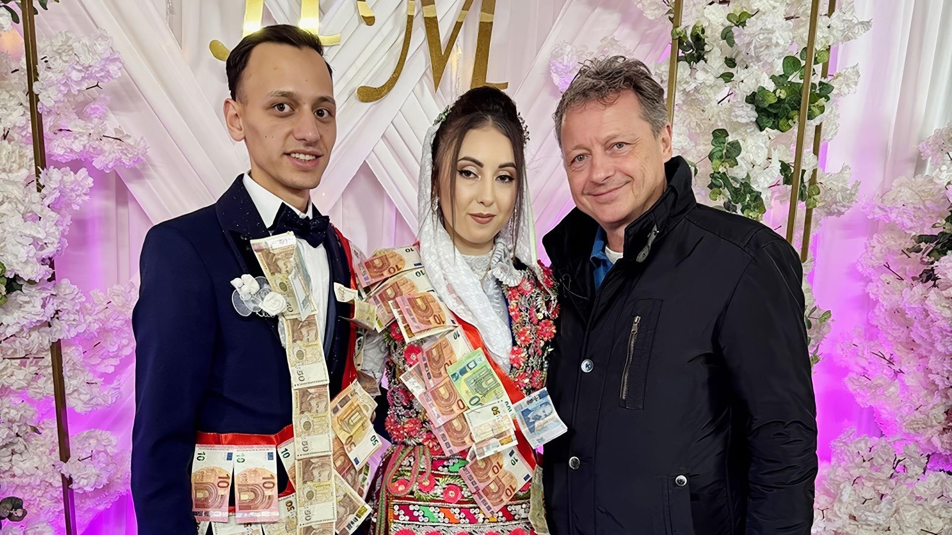 Das Bild zeigt eine Hochzeitsfeier in einem festlichen Moment. Zwei Personen, ein Bräutigam und eine Braut, stehen in der Mitte und lächeln. Der Bräutigam trägt einen eleganten Anzug mit einer Fliege und hat Geldscheine über die Brust drapiert. Die Braut trägt ein traditionelles, bunt verziertes Kleid mit Blumenmustern und hat ebenfalls Geldscheine an ihrer Kleidung angebracht. Sie trägt eine weiße Kopfbedeckung, die auf Hochzeitsbräuche hinweist.  Im Hintergrund ist eine dekorierte Kulisse mit großen, weißen Stoffbahnen und einem goldenen Schriftzug „HM“ sichtbar. Zudem sind künstliche Blumenarrangements zu sehen, die den Raum festlich schmücken. Die Umgebung hat eine farbige Beleuchtung, die dem Ambiente eine warme und feierliche Atmosphäre verleiht. Rechts von den beiden Hochzeitsgästen steht ein älterer Herr, der ebenfalls lächelt und in einem schwarzen Mantel gekleidet ist. Der Boden ist gefliest.   Das Bild vermittelt die fröhliche Stimmung einer traditionellen Hochzeit und zeigt die kulturellen Bräuche, die mit solchen Feierlichkeiten verbunden sind.