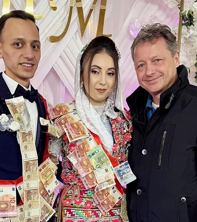 Das Bild zeigt eine Hochzeitsfeier in einem festlichen Moment. Zwei Personen, ein Bräutigam und eine Braut, stehen in der Mitte und lächeln. Der Bräutigam trägt einen eleganten Anzug mit einer Fliege und hat Geldscheine über die Brust drapiert. Die Braut trägt ein traditionelles, bunt verziertes Kleid mit Blumenmustern und hat ebenfalls Geldscheine an ihrer Kleidung angebracht. Sie trägt eine weiße Kopfbedeckung, die auf Hochzeitsbräuche hinweist.  Im Hintergrund ist eine dekorierte Kulisse mit großen, weißen Stoffbahnen und einem goldenen Schriftzug „HM“ sichtbar. Zudem sind künstliche Blumenarrangements zu sehen, die den Raum festlich schmücken. Die Umgebung hat eine farbige Beleuchtung, die dem Ambiente eine warme und feierliche Atmosphäre verleiht. Rechts von den beiden Hochzeitsgästen steht ein älterer Herr, der ebenfalls lächelt und in einem schwarzen Mantel gekleidet ist. Der Boden ist gefliest.   Das Bild vermittelt die fröhliche Stimmung einer traditionellen Hochzeit und zeigt die kulturellen Bräuche, die mit solchen Feierlichkeiten verbunden sind.