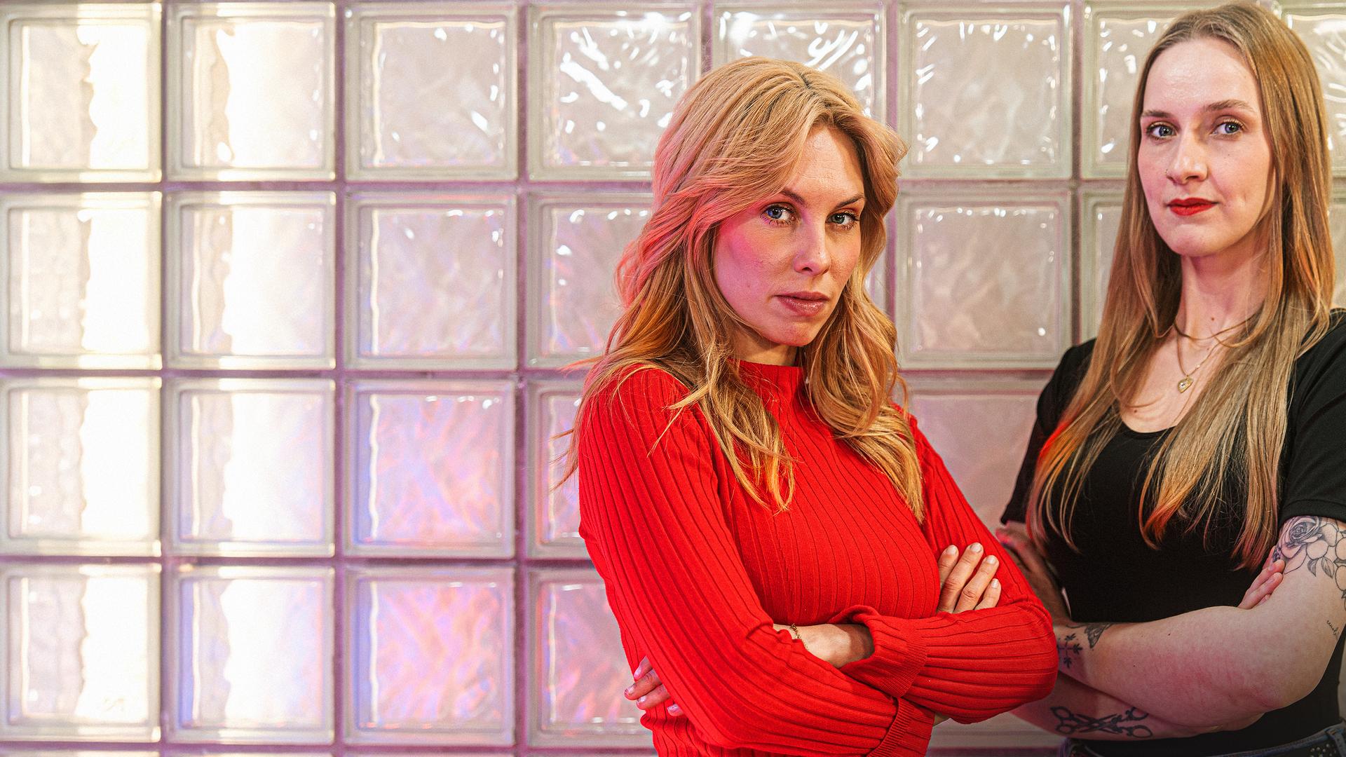 Zwei junge Frauen stehen mit verschränkten Armen und ernstem Blick vor einer Wand.