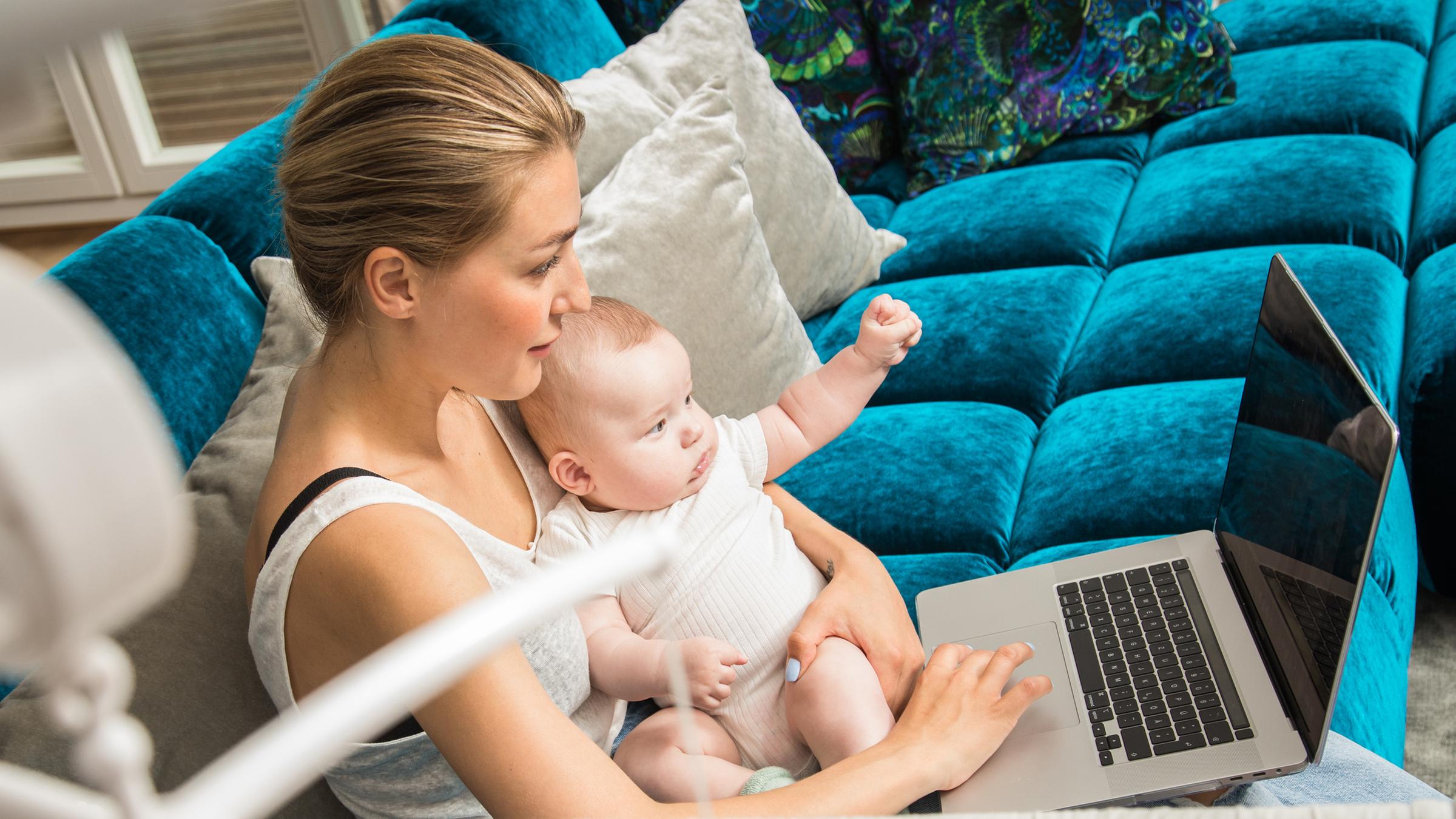 Eine Frau hält am 24.06.2021 in Berlin ihr Baby auf dem Schoss, waehrend sie an einem Laptop arbeitet 