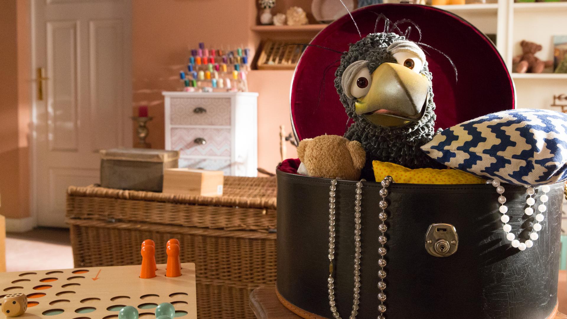 Rudi hockt in seiner Hutschachtel und schaut neugierig in den Laden hinüber. Neben ihm steht ein aufgebautes Brett-Spiel.