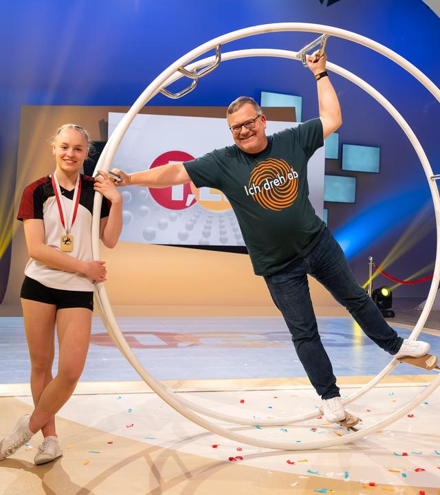  Junioren-Weltmeisterin Lea Gmeiner lehnt lässig an einem Rhönrad. Elton hält sich im Rhönrad mit den Händen fest und lächelt in die Kamera.