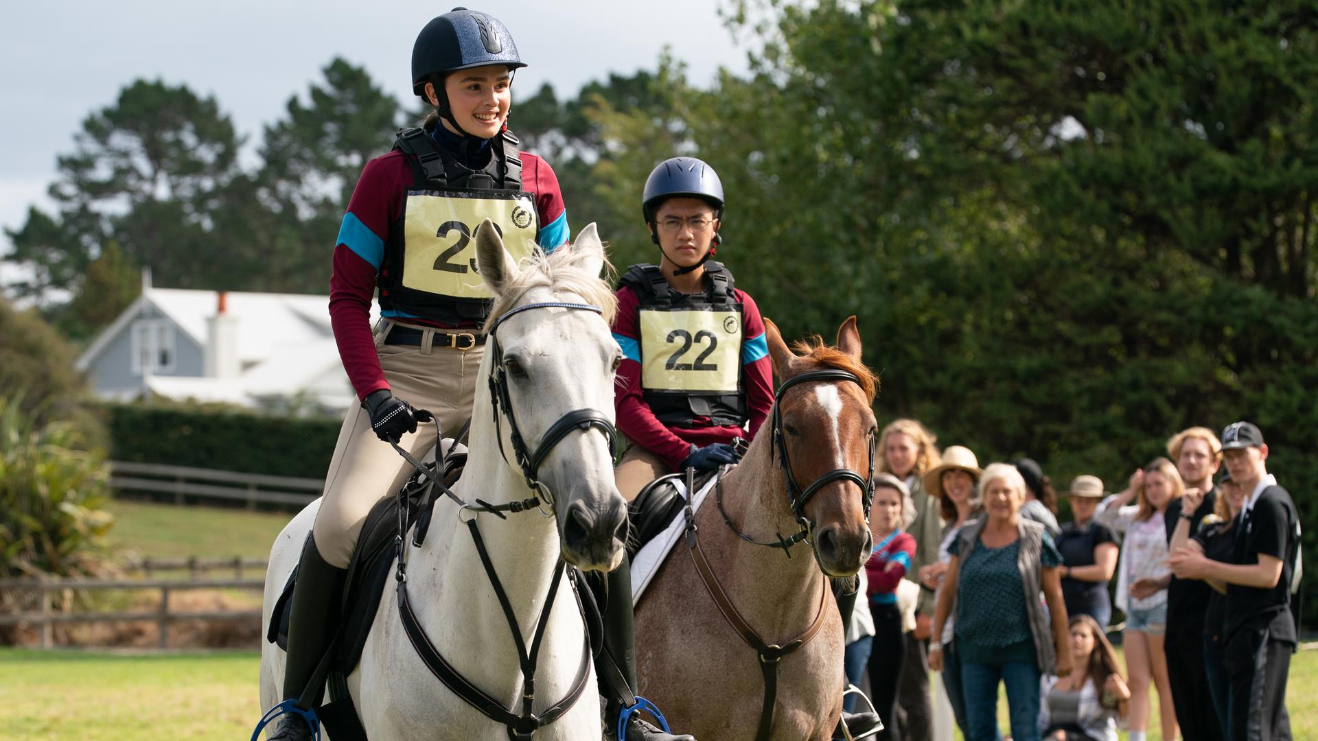Natasha und Caleb reiten ihre Pferde beim Turnier gegen die Rivalinnen von Dulmoth Park. Beide tragen Helm, Schutzweste und ihre Startnummern. Im Hintergrund stehen einige Zuschauerinnen und Zuschauer.
