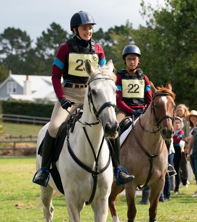 Natasha und Caleb reiten ihre Pferde beim Turnier gegen die Rivalinnen von Dulmoth Park. Beide tragen Helm, Schutzweste und ihre Startnummern. Im Hintergrund stehen einige Zuschauerinnen und Zuschauer.