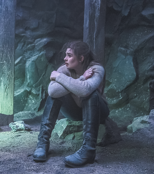 Natascha sitzt in einer dunklen Höhle zusammengekauert auf einem Stein und blickt nach links, wo ein Lichtstrahl hinfällt. Im Hintergrund sind Stützbalken zu sehen.