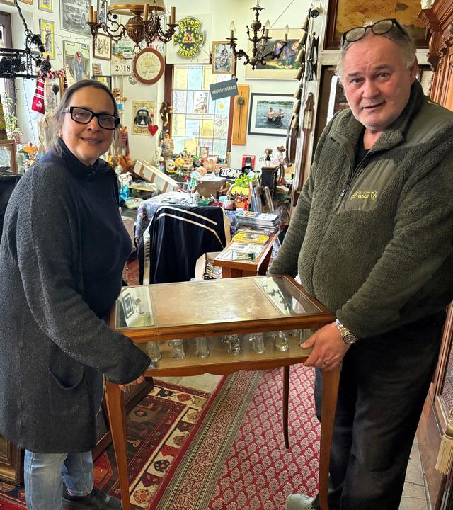 Sonja und Waldi (v.l.n.r.) stehen in einem Antiquitätenladen und halten einen alten Beistelltisch in den Händen und lächeln in die Kamera. 