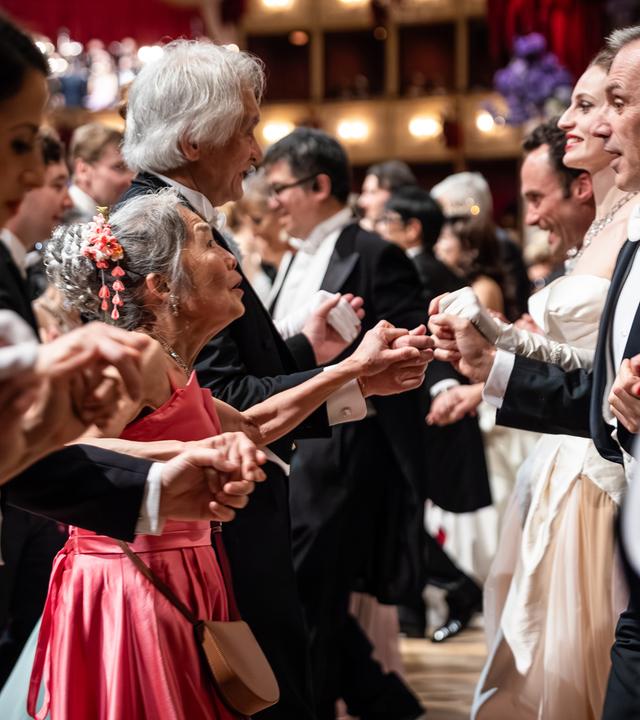 Das Bild zeigt eine Tanzszene während des Opernballs 2026. In einem festlich dekorierten Saal, der elegant mit roten Vorhängen und Kristallleuchtern ausgestattet ist, befinden sich zahlreiche Ballgäste, die in Paaren tanzen.   Im Vordergrund sind mehrere Personen sichtbar, die sich an den Händen halten. Eine ältere Frau mit grauen Haaren und einer pinken, fließenden Kleid sieht fröhlich aus. Sie tanzt neben einer Frau in einem weißen, eleganten Kleid. Um sie herum sind auch andere Gäste in formeller Abendkleidung, darunter Herren in Smoking und Damen in formellen Ballkleidern.  Der Hintergrund zeigt eine Mischung aus weiteren Tänzern und einem erhöhten Bereich, wahrscheinlich eine Bühne, auf der möglicherweise eine musikalische Darbietung stattfindet. Der gesamte Raum vermittelt eine festliche und glamouröse Atmosphäre des Balls.