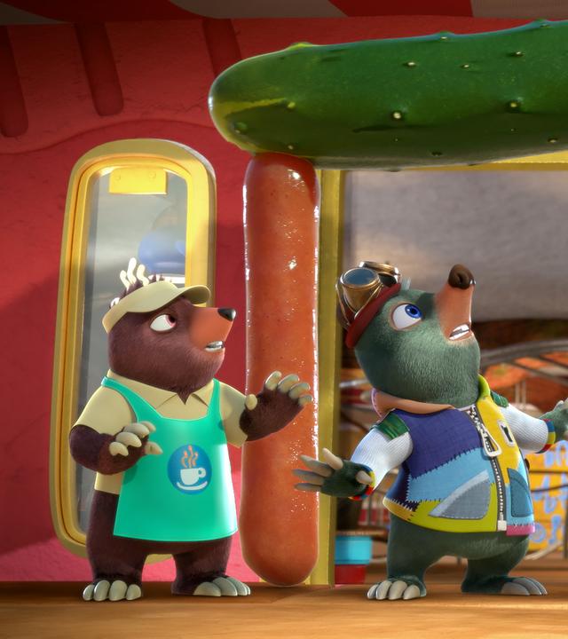 Moley steht im Eingang des Cafés und blickt senkrecht nach oben auf eine große Salatgurke, die gemeinsam mit zwei riesigen Würstchen den Türsturz und die Säulen des Eingangs bildet. Neben ihm sind einige andere Maulwürfe zu sehen.