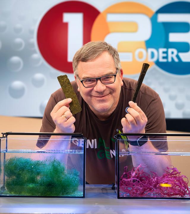 Elton lehnt sich über zwei kleine Aquarien in denen unterschiedliche Algenarten zu sehen sind. In der Hand hält Nudeln und Schokolade aus Algen.