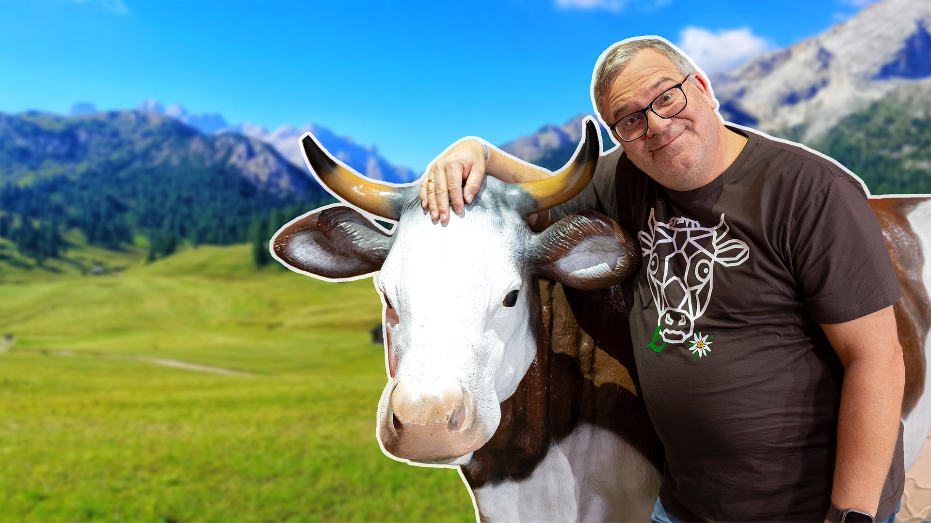 Elton steht vor einer Plastikkuh im Alpen-Panorama
