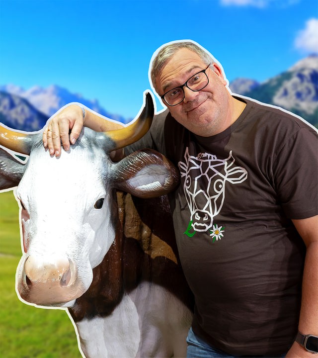 Elton steht vor einer Plastikkuh im Alpen-Panorama