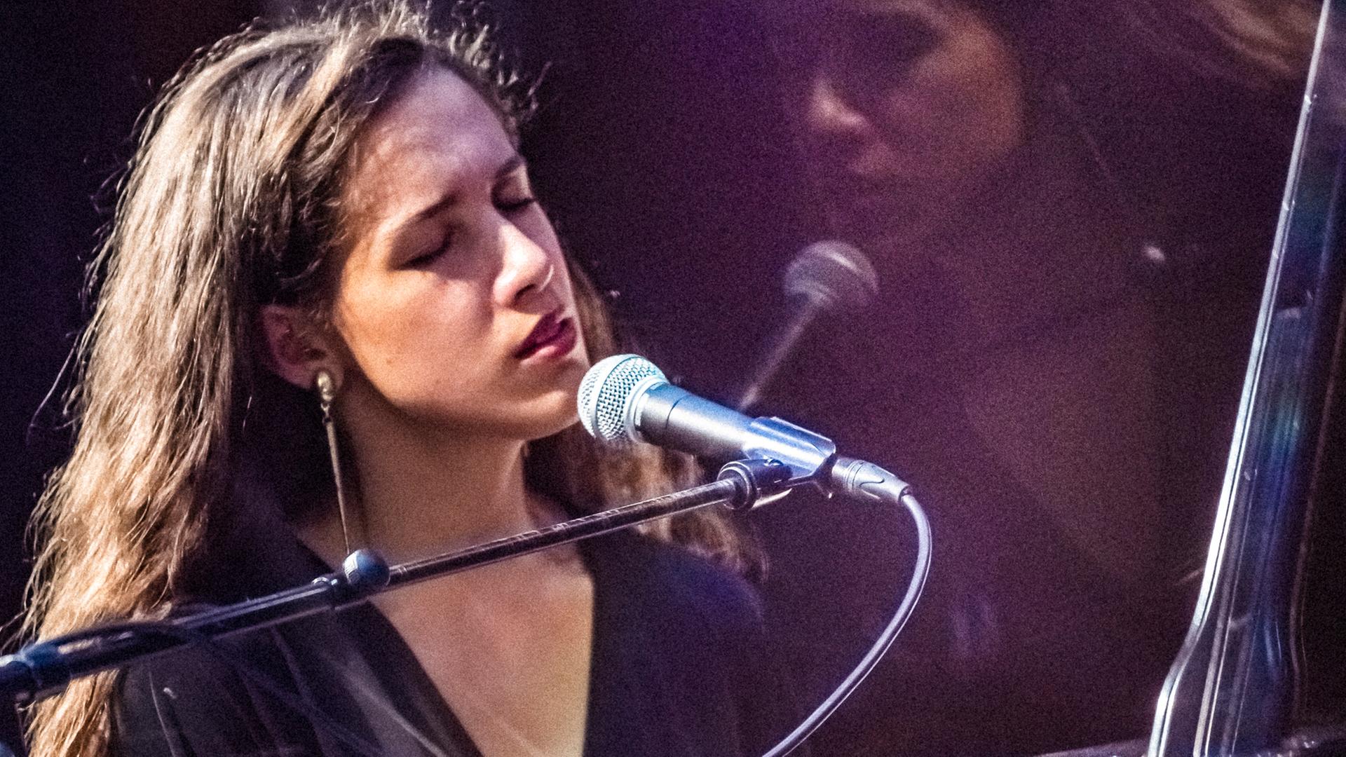Musizierende Person am Mikrofon auf einer Bühne, beleuchtet von Bühnenlicht, mit sichtbarem Instrument und Performance-Situation.
