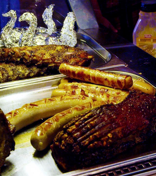 Das Bild zeigt eine Theke mit verschiedenen Grillgerichten. Im Vordergrund sind mehrere Fleischstücke und Würstchen auf einem rechteckigen Edelstahlblech angeordnet.   - Von links nach rechts sind sichtbar:   - Ein großes Stück Fleisch, wahrscheinlich Schweinebraten, mit einer glänzenden, leicht knusprigen Oberfläche und Rillen.   - Daneben liegt ein Rippchen, ebenfalls mit einer knusprigen Kruste.   - Vor dem Rippchen befinden sich mehrere Würstchen in verschiedenen Größen und Formen, einige scheinen gegrillt zu sein.    Hinter den Fleischgerichten sind zwei dekorative Schwäne aus Alufolie sichtbar, die die Präsentation auflockern. Im Hintergrund sind einige Flaschen mit gelben und weißen Flüssigkeiten zu erkennen, möglicherweise Saucen oder Dressing.  Die gesamte Szenerie wirkt einladend und typisch für eine gastronomische Einrichtung in den Bergen, wie sie in der Alpengastronomie zu finden ist.