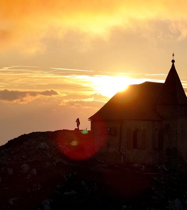 Das Bild zeigt die Dobratsch Deutsche Kapelle, eine Kirche auf einem Berggipfel in den Alpen. Die Kapelle hat einen spitzen Turm und ist im Hintergrund vor einem dramatischen Sonnenuntergang positioniert, bei dem orangefarbene und goldene Töne den Himmel füllen. Der Sonnenstrahl scheint hinter der Kapelle hervor und erzeugt eine leuchtende Silhouette. Im Vordergrund ist ein felsiger Hang zu sehen, und auf diesem Hang steht eine Person, die möglicherweise die Aussicht genießt. Die umgebenden Bereiche sind von Wolken umgeben, was dem Bild eine mystische Atmosphäre verleiht.