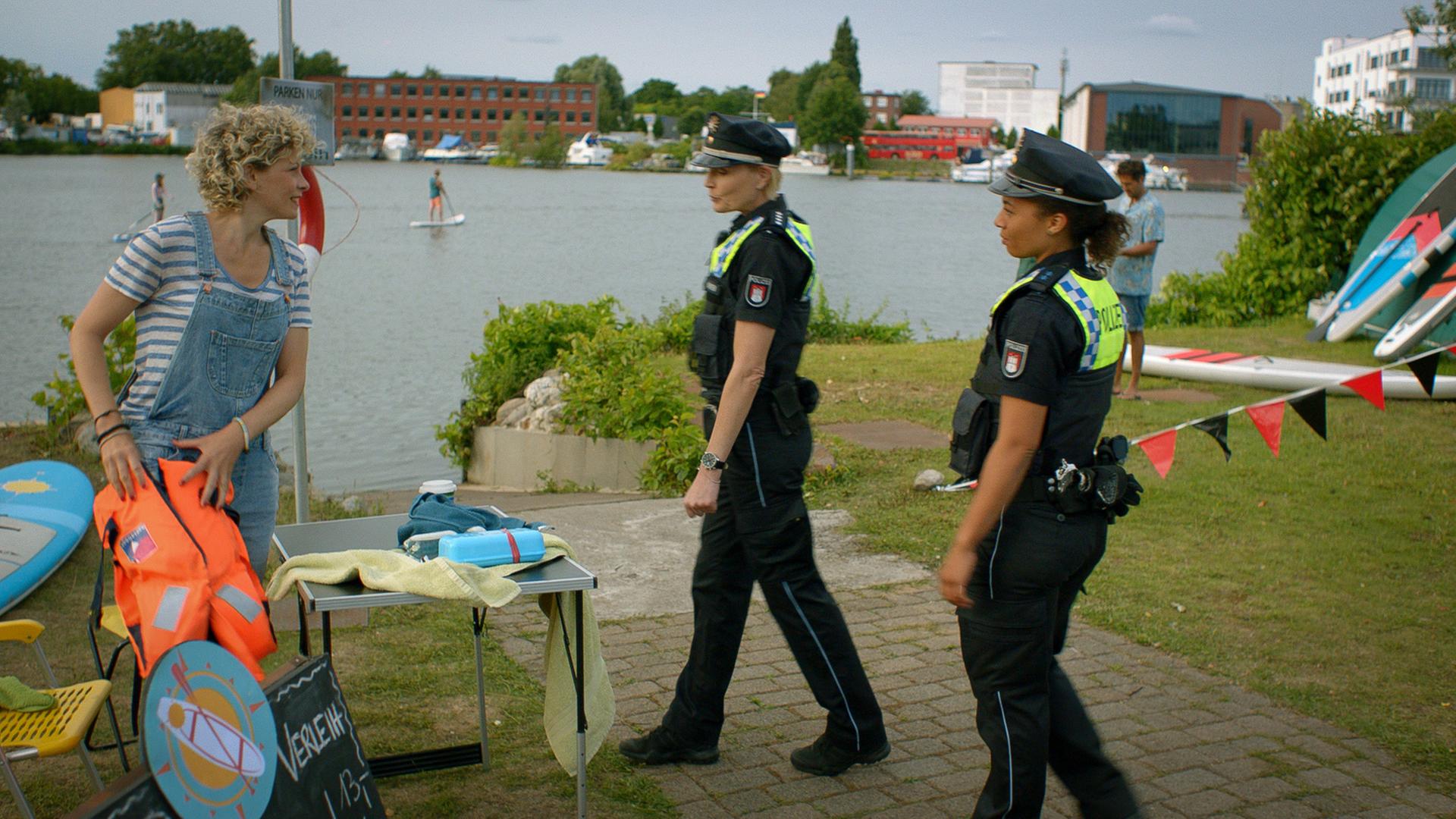 "Notruf Hafenkante - Alstervergnügen": Die Polizistinnen Melanie Hansen (Sanna Englund) und Daisy Petersen (Aysha Joy Samuel) stehen an einer Anlegestelle bei Viktoria Lamprecht (Anna Schäfer), die gerade im Begriff ist, eine Rettungsweste zusammenzufalten. Viktoria grinst zu den Polizistinnen.