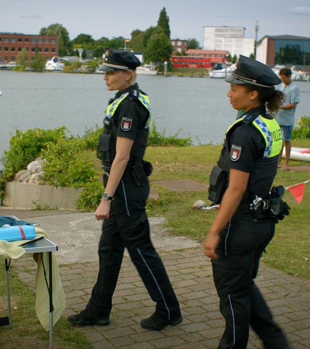 "Notruf Hafenkante - Alstervergnügen": Die Polizistinnen Melanie Hansen (Sanna Englund) und Daisy Petersen (Aysha Joy Samuel) stehen an einer Anlegestelle bei Viktoria Lamprecht (Anna Schäfer), die gerade im Begriff ist, eine Rettungsweste zusammenzufalten. Viktoria grinst zu den Polizistinnen.
