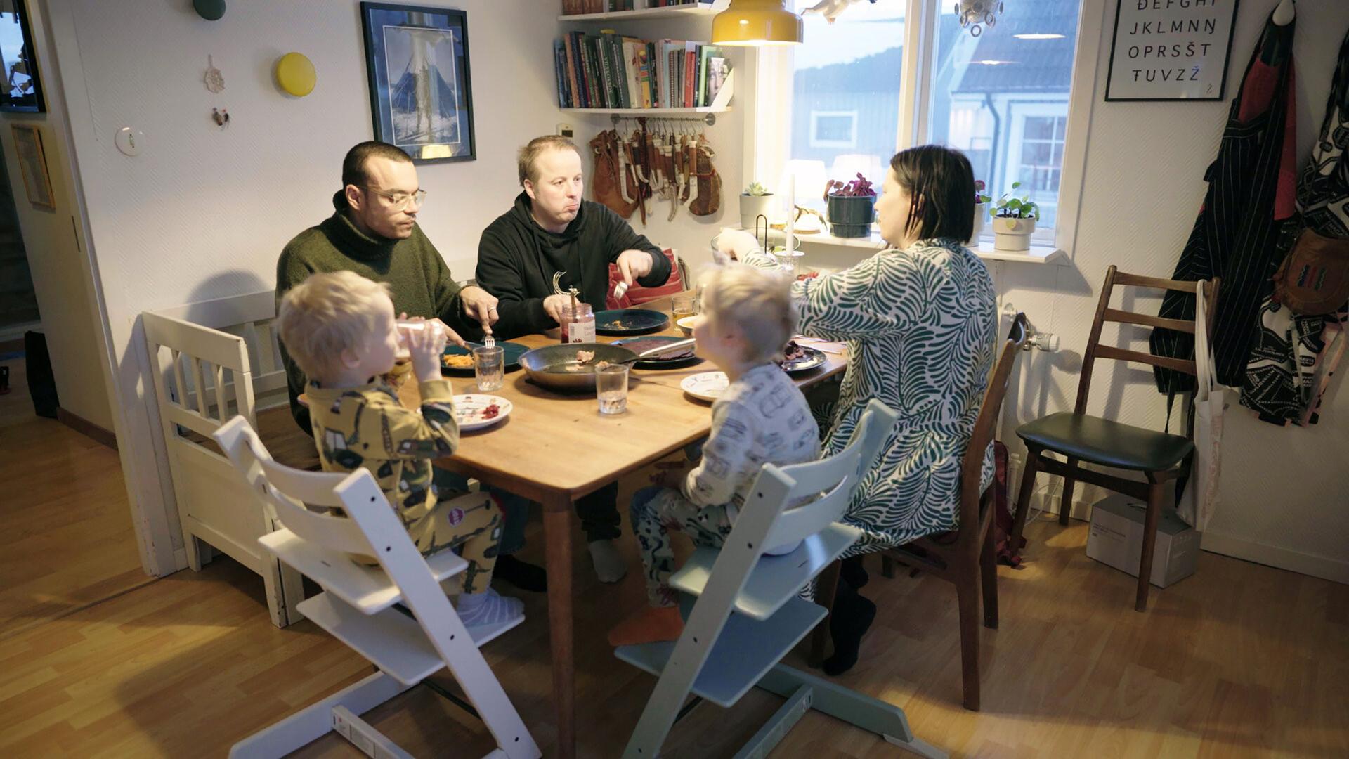 Das Bild zeigt eine familiäre Essenssituation in einem gemütlichen Innenraum. An einem rechteckigen Holztisch sitzen fünf Personen. Zwei Erwachsene und drei Kinder sind um den Tisch arrangiert.   Die Erwachsenen sind mittleren Alters, einer trägt einen grünen Pullover und eine Brille, der andere einen schwarzen Hoodie. Die Frauen sind unterschiedlich gekleidet; eine trägt ein Kleid mit einem blauen und weißen Muster. Die Kinder sind in Schlafanzügen gekleidet, einer in Gelb mit Maschinenmotiven, ein anderer in Weiß mit bunten Punkten.  Auf dem Tisch sind mehrere Teller mit Essen, Trinkgläser und eine offene Glasflasche mit Konfitüre. Im Hintergrund sind Regale mit Büchern und einigen dekorativen Elementen zu sehen. Ein Fenster an der Wand zeigt eine ländliche Aussicht auf die schwedische Arktis.  Die Umgebung wirkt einladend und warm, mit Holzfußboden und sanfter Beleuchtung durch eine Lampe über dem Tisch.