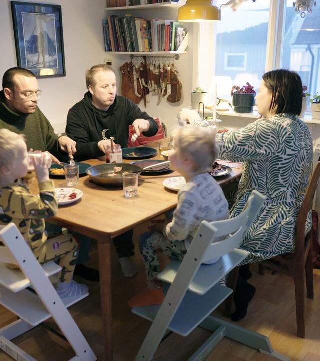 Das Bild zeigt eine familiäre Essenssituation in einem gemütlichen Innenraum. An einem rechteckigen Holztisch sitzen fünf Personen. Zwei Erwachsene und drei Kinder sind um den Tisch arrangiert.   Die Erwachsenen sind mittleren Alters, einer trägt einen grünen Pullover und eine Brille, der andere einen schwarzen Hoodie. Die Frauen sind unterschiedlich gekleidet; eine trägt ein Kleid mit einem blauen und weißen Muster. Die Kinder sind in Schlafanzügen gekleidet, einer in Gelb mit Maschinenmotiven, ein anderer in Weiß mit bunten Punkten.  Auf dem Tisch sind mehrere Teller mit Essen, Trinkgläser und eine offene Glasflasche mit Konfitüre. Im Hintergrund sind Regale mit Büchern und einigen dekorativen Elementen zu sehen. Ein Fenster an der Wand zeigt eine ländliche Aussicht auf die schwedische Arktis.  Die Umgebung wirkt einladend und warm, mit Holzfußboden und sanfter Beleuchtung durch eine Lampe über dem Tisch.