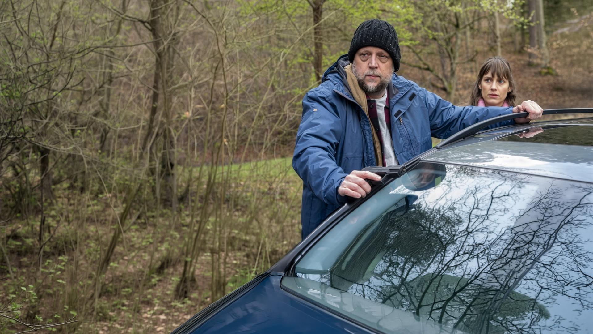 "SOKO Stuttgart - Alte Schulden": Philip Töpfer (Smudo) steht mit einer Pistole in der Hand am Waldrand und lehnt sich mit beiden Händen gegen ein Auto. Hinter ihm steht Anne Dorn (Sophie Lutz). Sie schaut ihn an, während er nachdenklich in die Ferne blickt.