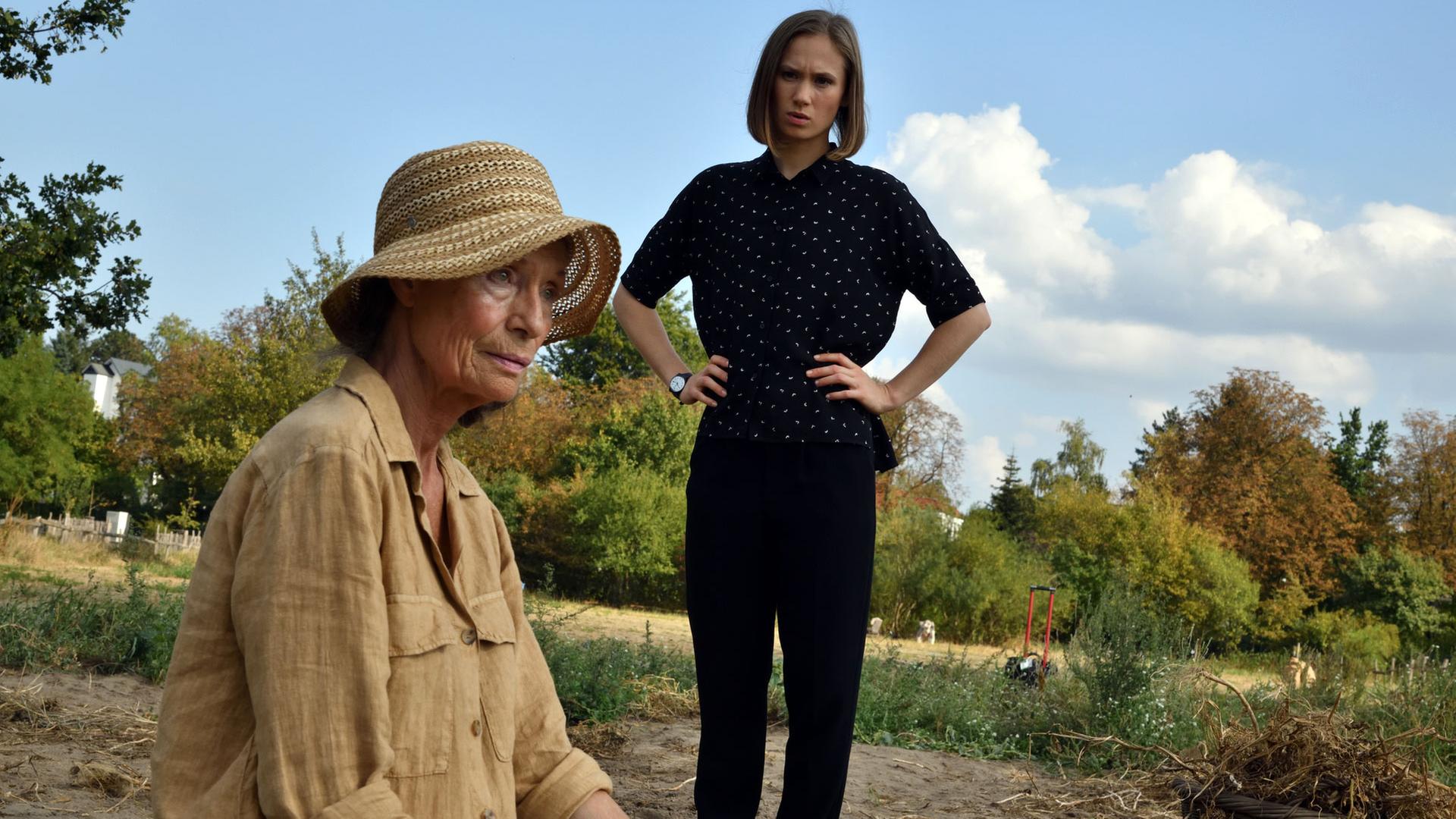 "Die Spezialisten - Alte Wunden": Dr. Julia Löwe (Alina Levshin) steht in einem Feld. Sie hat die Hände in die Hüften gestemmt und blickt nachdenklich zu Anna Rivolta (Renate Serwotke), die neben ihr in der Hocke auf dem Boden sitzt. Neben ihr steht ein Weidenkorb. Sie starrt gedankenverloren vor sich ins Leere.