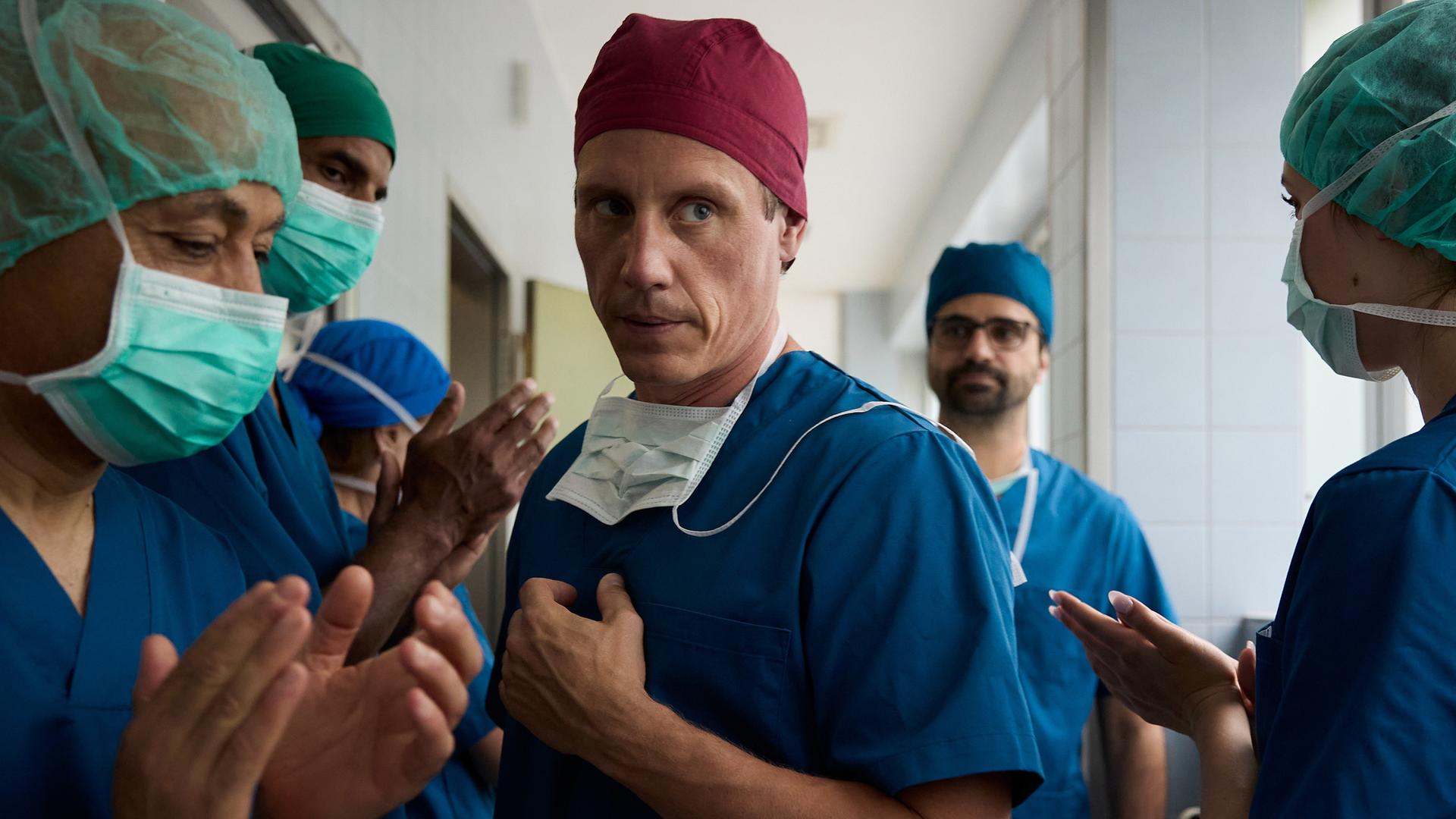  Im Flur der Klinik läuft Dr. Nice (Patrick Kalupa) an seinen applaudierenden Mitarbeitern vorbei. Dr. Schmidtke (Maximilian Grill) läuft etwas abseits hinter ihm her und beobachtet dies missmutig.