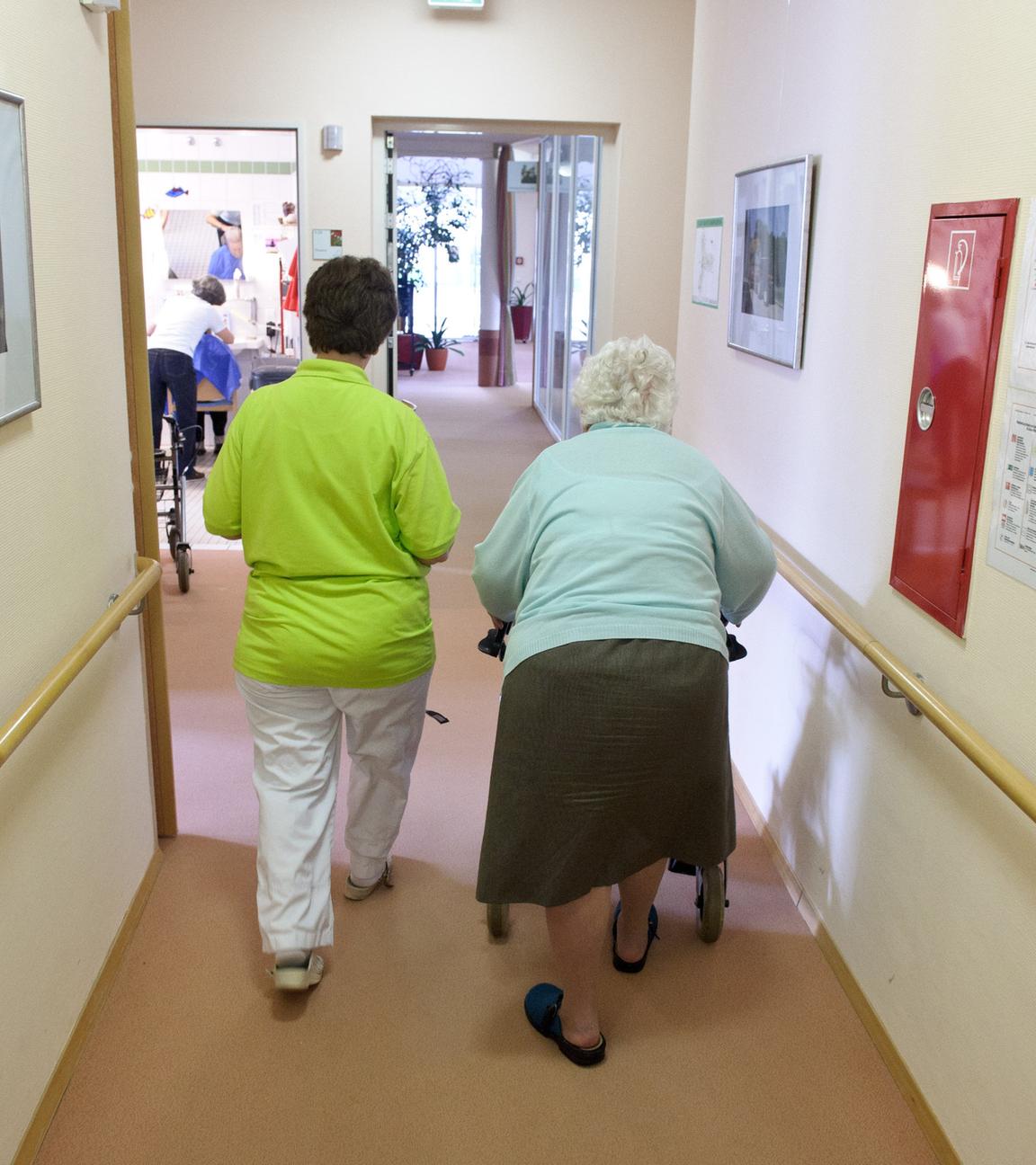 Eine Altenpflegerin begleitet eine Seniorin an einem Rollator durch einen Flur.