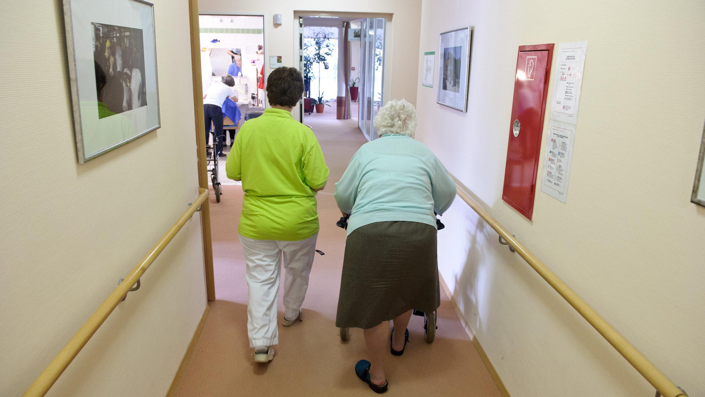 Eine Altenpflegerin begleitet eine Seniorin an einem Rollator durch einen Flur.