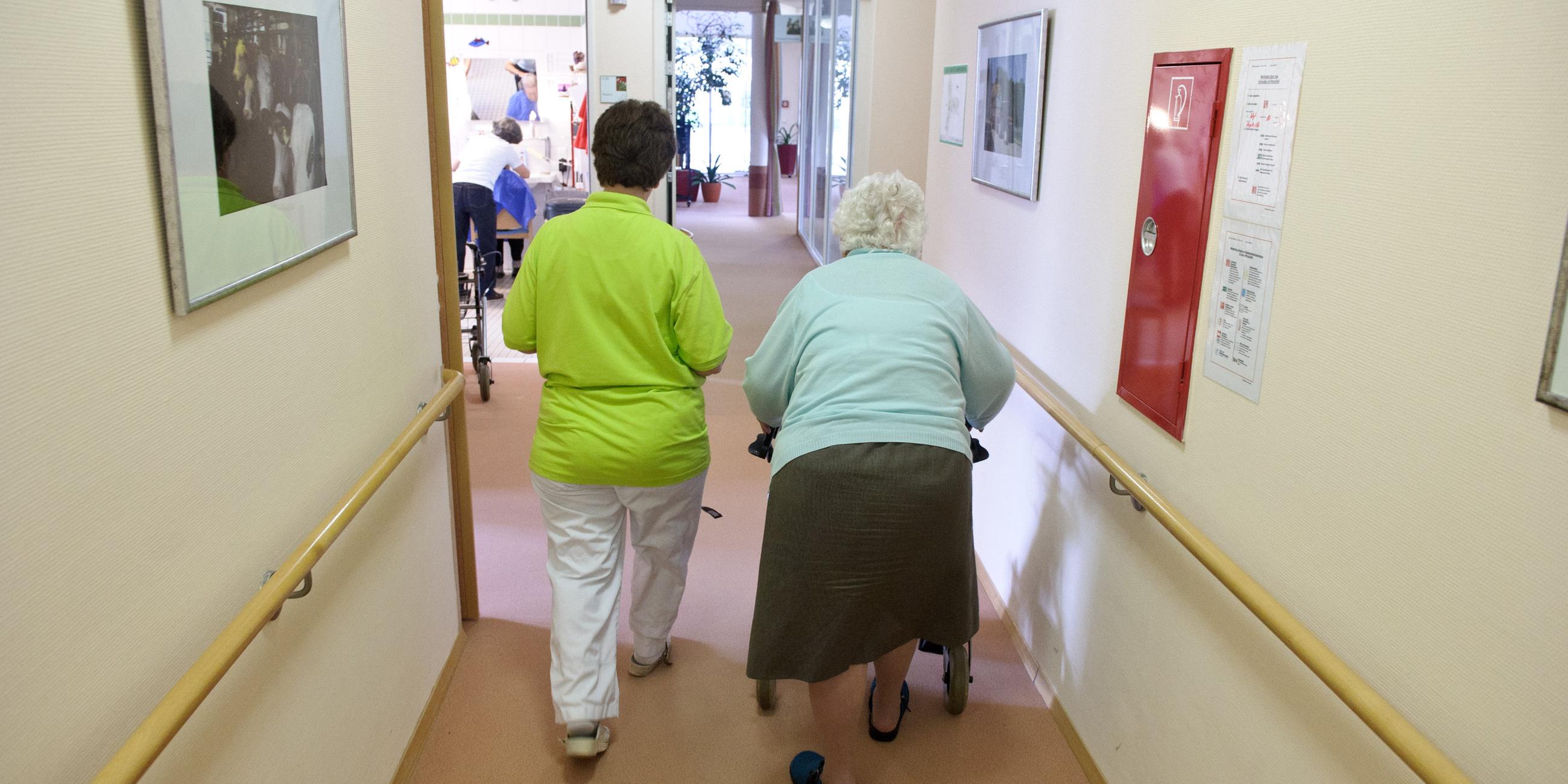 Eine Altenpflegerin begleitet eine Seniorin an einem Rollator durch einen Flur.
