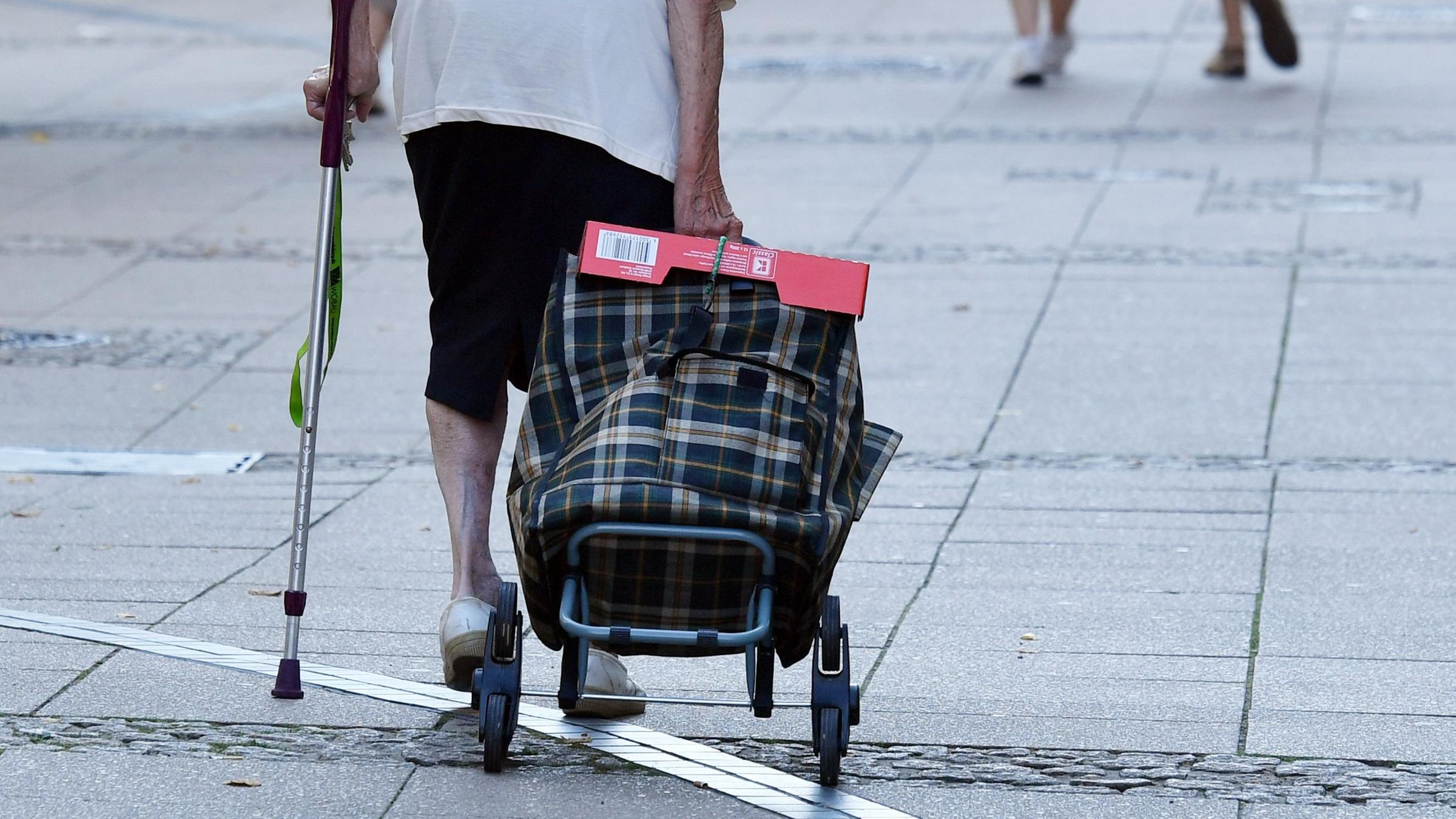 Eine alte Frau mit einer Krücke und Einkaufstrolley geht durch eine Fußgängerstraße.
