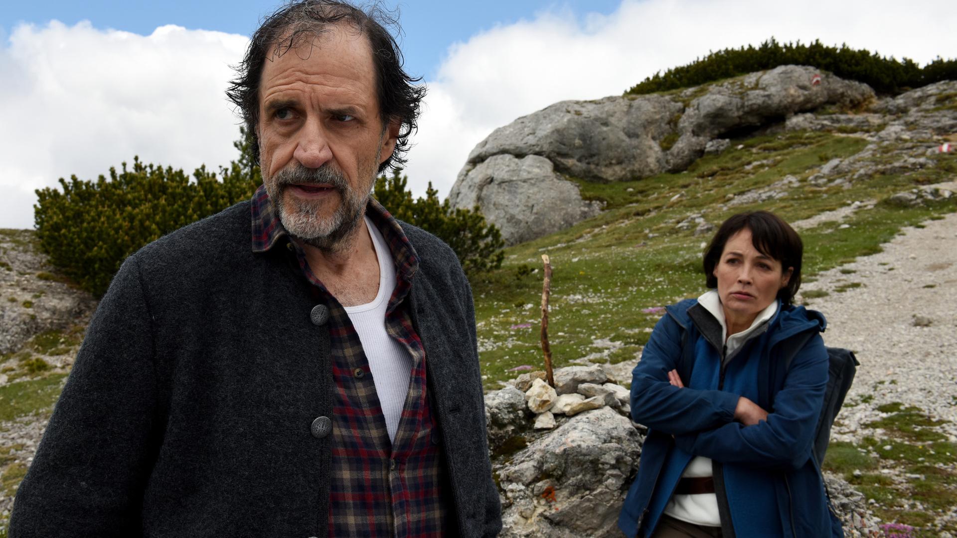 "Die Bergretter - Altes Eisen": Tabea Gollini (Anke Sevenich) lehnt mit beleidigter Miene und verschränkten Armen an einem Stein, der an einem Berghang liegt. Vor ihr steht abgewandt Walther Senn (Helmfried von Lüttichau) und blickt genervt in die Ferne.