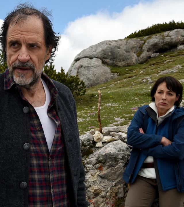 "Die Bergretter - Altes Eisen": Tabea Gollini (Anke Sevenich) lehnt mit beleidigter Miene und verschränkten Armen an einem Stein, der an einem Berghang liegt. Vor ihr steht abgewandt Walther Senn (Helmfried von Lüttichau) und blickt genervt in die Ferne.