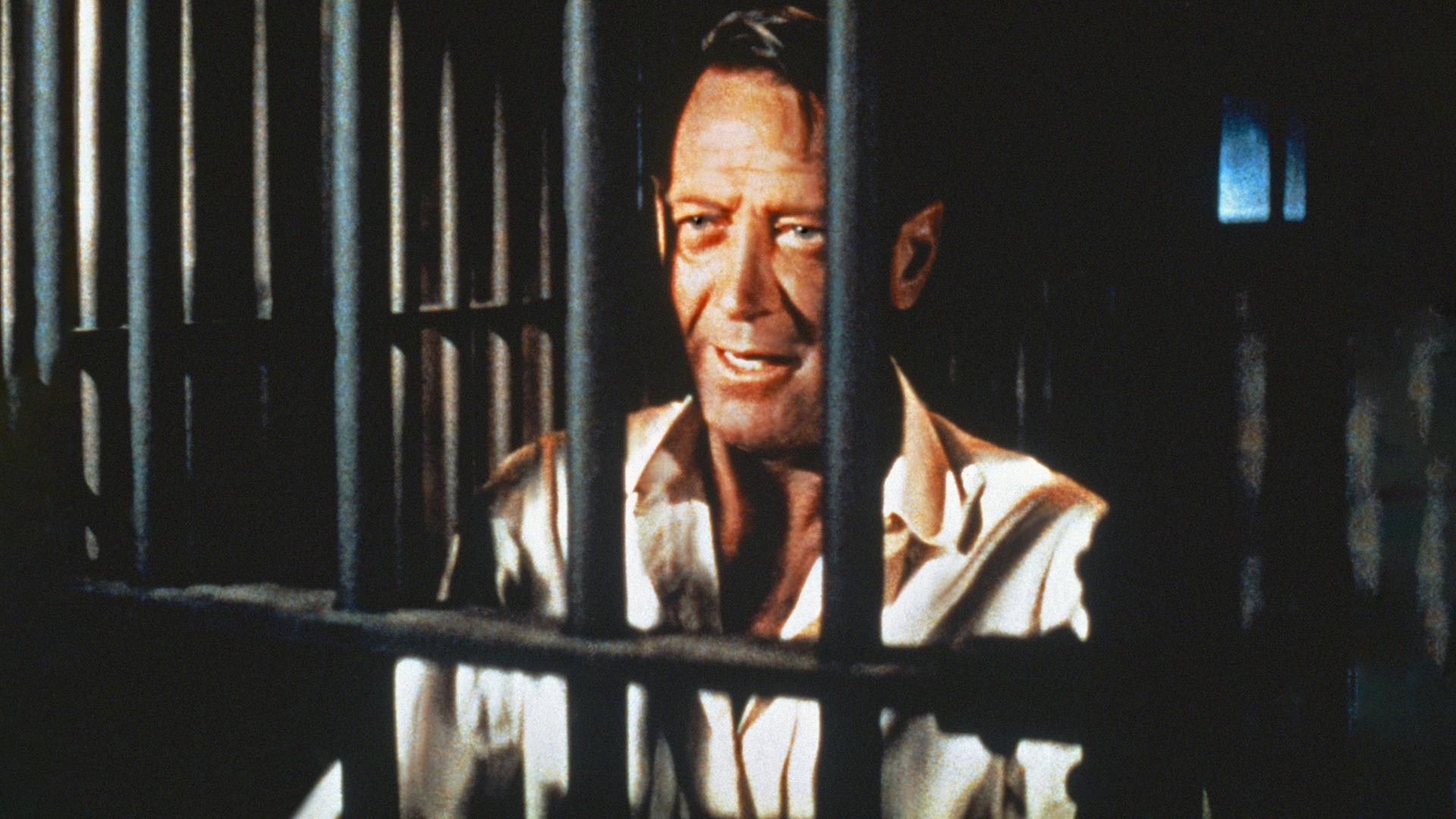 Alvarez Kelly ist mit weißem Hemd im Close-Up hinter schwarzen Gefängnisgittern zu sehen. Trotz seiner Situation und Zeichen der Müdigkeit im Gesicht lächelt er.