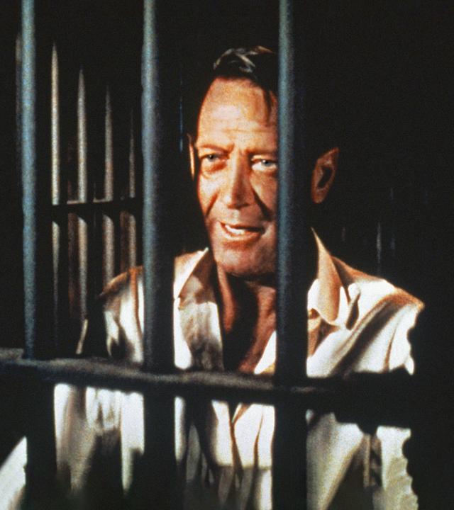 Alvarez Kelly ist mit weißem Hemd im Close-Up hinter schwarzen Gefängnisgittern zu sehen. Trotz seiner Situation und Zeichen der Müdigkeit im Gesicht lächelt er.