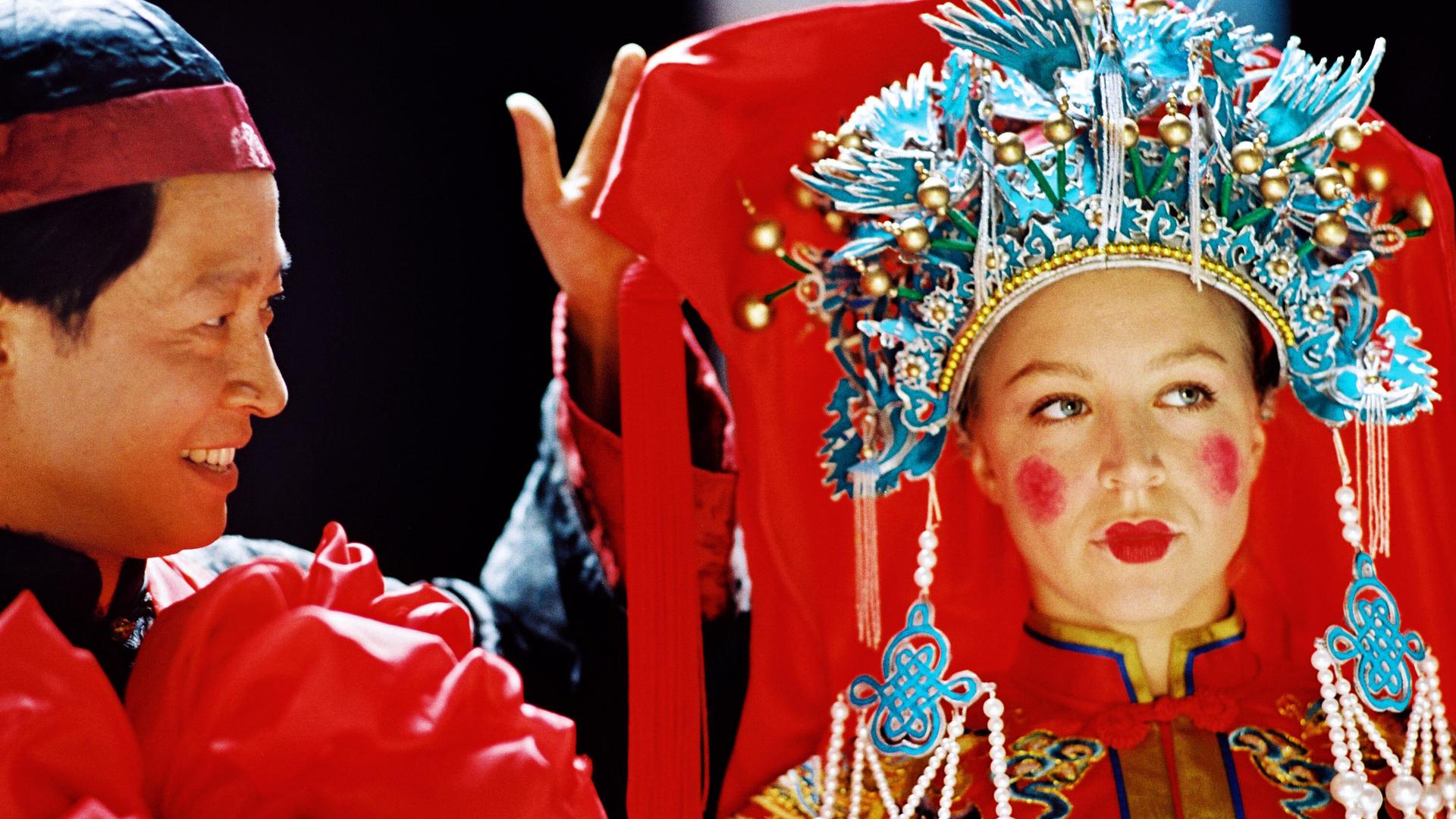 Eine junge Frau in aufwendigem, traditionell chinesischem Brautgewand mit perlenbesetztem Kopfschmuck steht neben einem lächelnden Mann in festlicher Kleidung